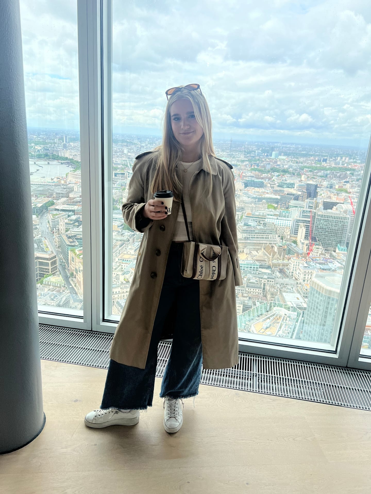 Carson Miller in a trench coat at a high window overlooking the London skyline.